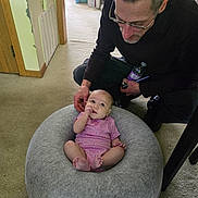 Kaislee is registered to the contest to win money with this photo: baby, infant, man, adult, glasses, pink_onesie, cushion, carpet, indoor, bottle, person, floor, home, curious, seated, looking_down, casual_clothing, light, furniture, doorway