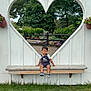 toddler, child, bench, heart_shape, flower_pots, greenery, grass, outdoor, smiling, cute, white_wall, wood, plants, nature, daylight, casual_clothing, sneakers, park, fence, playful