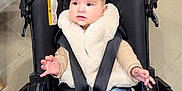Cynthia is registered to the contest to win money with this photo: baby, barefoot, black_stroller, child, clothing, colorful, cute, expression, face, floor, foot, hands, indoor, infant, jeans, person, seatbelt, stroller, toy, vest