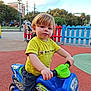 toddler, child, toy_bike, playground, yellow_shirt, blue_bike, shorts, outdoor, fence, stroller, trees, sky, curly_hair, play, summer, park, person, daylight, happy, cute