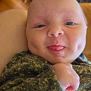 Noah is registered to the contest to win money with this photo: baby, infant, face, dimples, tongue, camouflage_clothing, hand, skin, head, person, indoor, furniture, chair, wood_floor, couch, cozy, expression, cute, portrait, newborn