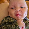 baby, infant, face, dimples, tongue, camouflage_clothing, hand, skin, head, person, indoor, furniture, chair, wood_floor, couch, cozy, expression, cute, portrait, newborn