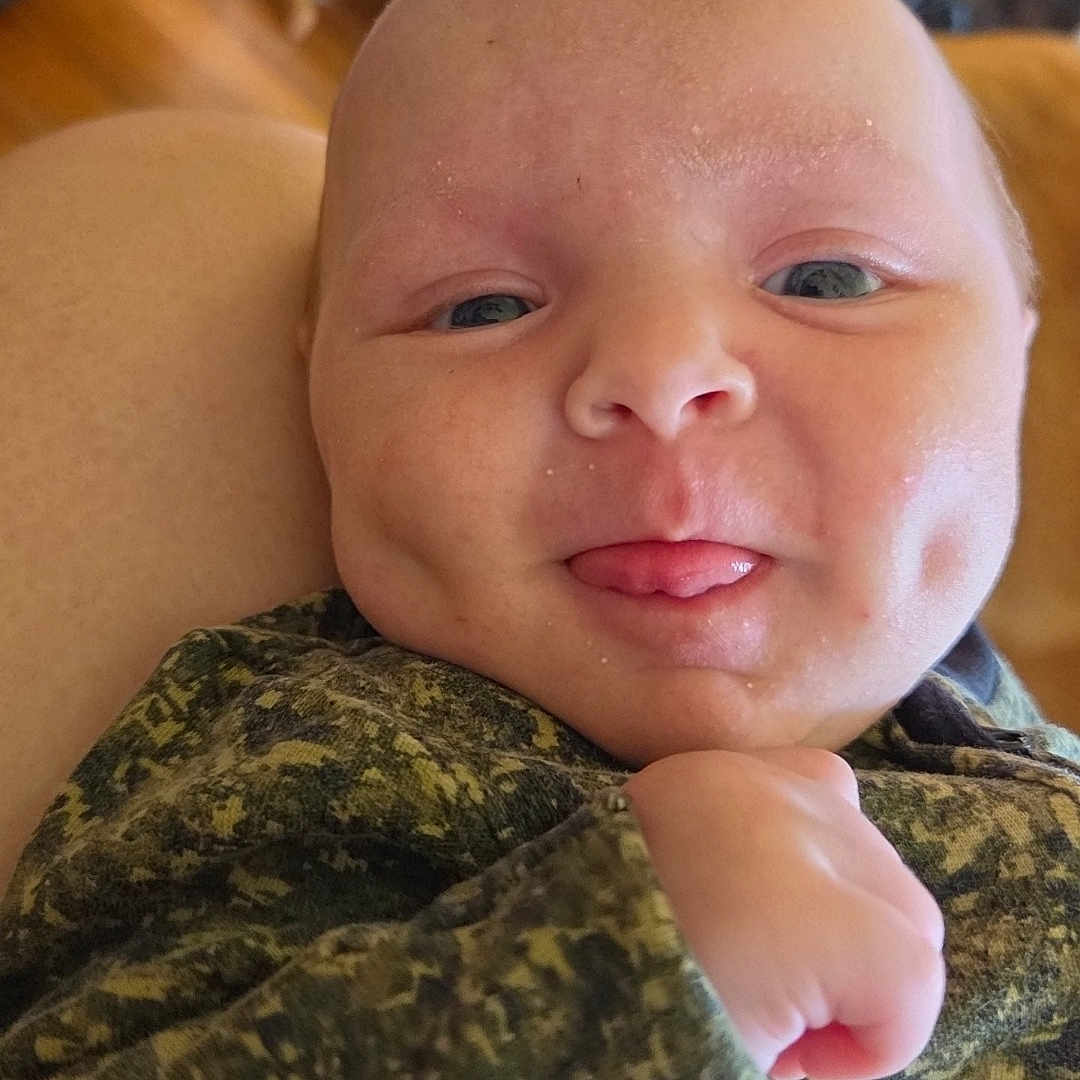 Noah is registered to the contest to win money with this photo: baby, infant, face, dimples, tongue, camouflage_clothing, hand, skin, head, person, indoor, furniture, chair, wood_floor, couch, cozy, expression, cute, portrait, newborn