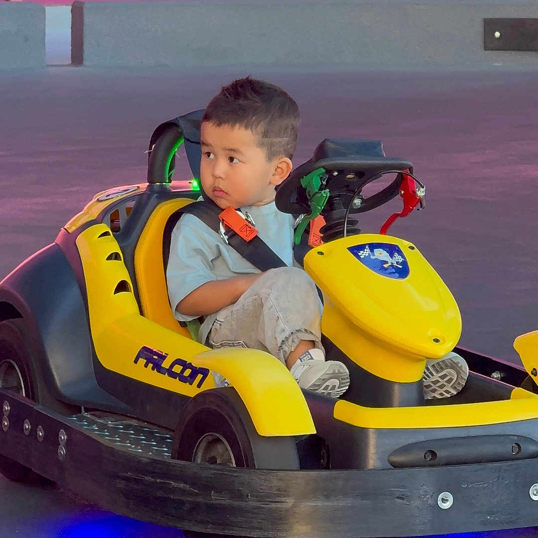 Dias joined the competition — help win amazing prizes! amusement_ride, arcade, bumper, candid, checkered_floor, child, colorful, face, go_kart, indoor, kid, neon_light, portrait, seat_belt, short_hair, sitting, sneakers, steering_wheel, wheel, yellow_vehicle