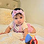 baby, child, pink_headband, pink_outfit, toy, bed, white_bedspread, pillow, indoor, cute, infant, person, reaching, soft_lighting, portrait, toddler, colorful_toy, headband, bedroom, crawling