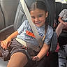 Keniyah is registered to the contest to win money with this photo: child, car_seat, seatbelt, phone, smile, braid, tshirt, shorts, vehicle_interior, window, road, sunlight, happy, kid, casual_clothing, seat, hand, portrait, travel, family