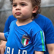 Giuseppe is registered to the contest to win money with this photo: toddler, child, curly_hair, blue_jersey, italia, soccer, sportswear, portrait, outdoor, person, face, expression, closeup, casual_clothing, young_child, short_sleeves, focused, background_blur, daylight, human