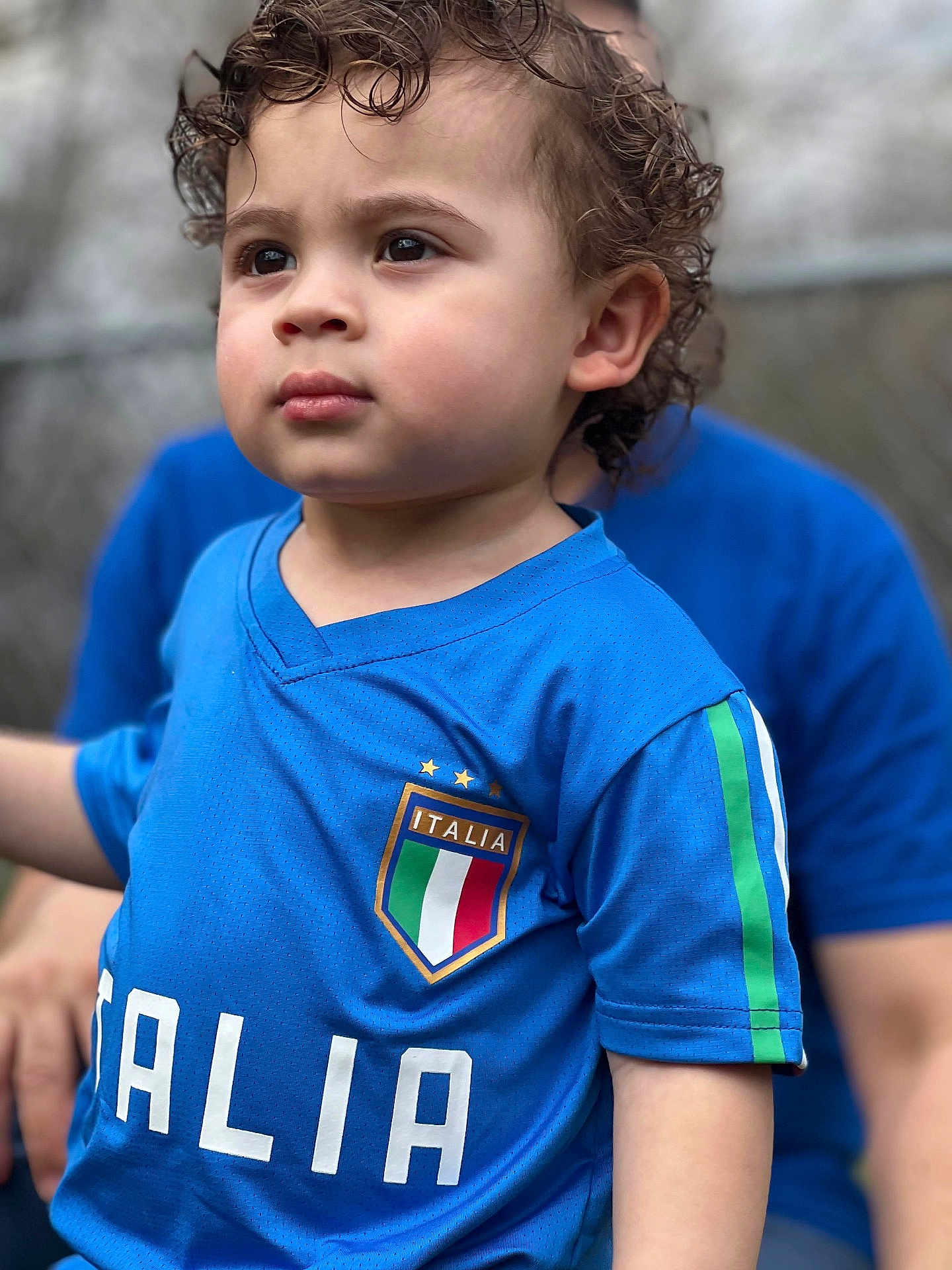 Giuseppe is registered to the contest to win money with this photo: toddler, child, curly_hair, blue_jersey, italia, soccer, sportswear, portrait, outdoor, person, face, expression, closeup, casual_clothing, young_child, short_sleeves, focused, background_blur, daylight, human