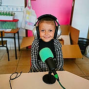 Loriana a rejoint le concours — aidez-le/la à gagner de superbes lots ! child, headphones, microphone, smiling, indoor, table, pink_door, cardboard_box, floor_tiles, black_and_white_clothing, person, happy, technology, audio, recording, cable, chair, furniture, room, portrait