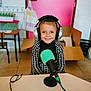 child, headphones, microphone, smiling, indoor, table, pink_door, cardboard_box, floor_tiles, black_and_white_clothing, person, happy, technology, audio, recording, cable, chair, furniture, room, portrait