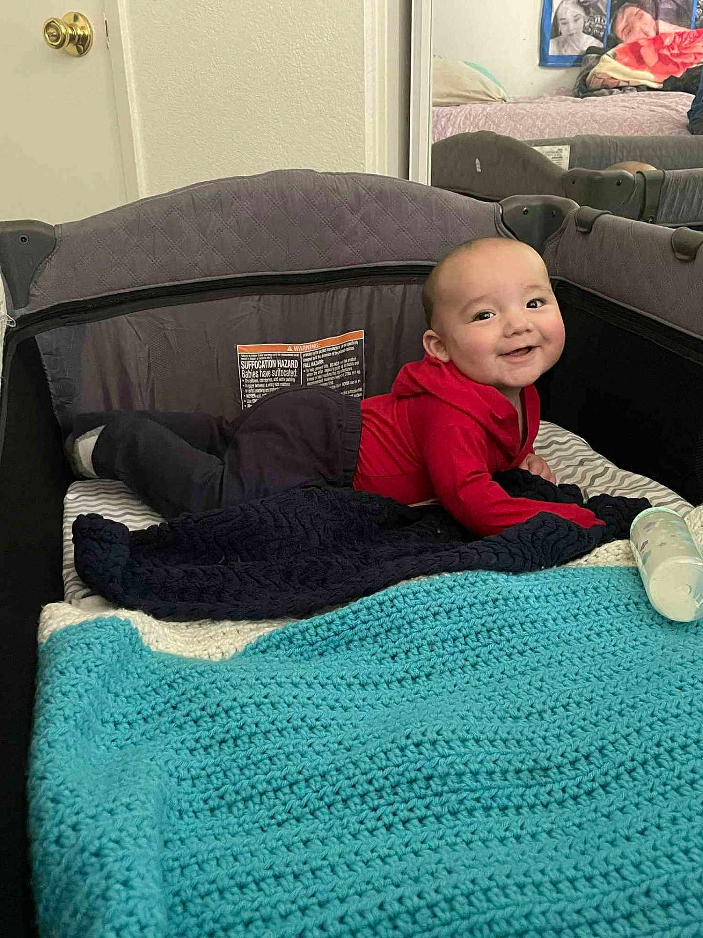 Alejandro is registered to the contest to win money with this photo: baby, playpen, blanket, red_hoodie, smiling, lying_down, baby_bottle, mirror, indoor, cozy, child, toddler, black_pants, fabric, cushion, home, happy, person, face, wall