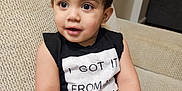 Adrian is registered to the contest to win money with this photo: toddler, child, couch, indoor, black_clothing, sleeveless_shirt, text_on_clothing, curly_hair, cute, portrait, seated, beige_background, happy, small_hands, big_eyes, casual, home, soft_lighting, close_up, young
