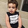 toddler, child, couch, indoor, black_clothing, sleeveless_shirt, text_on_clothing, curly_hair, cute, portrait, seated, beige_background, happy, small_hands, big_eyes, casual, home, soft_lighting, close_up, young
