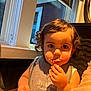 child, toddler, curly_hair, eating, food, bib, face, indoor, window, table, high_chair, curiosity, messy, portrait, person, baby, snack, cute, young_child, casual