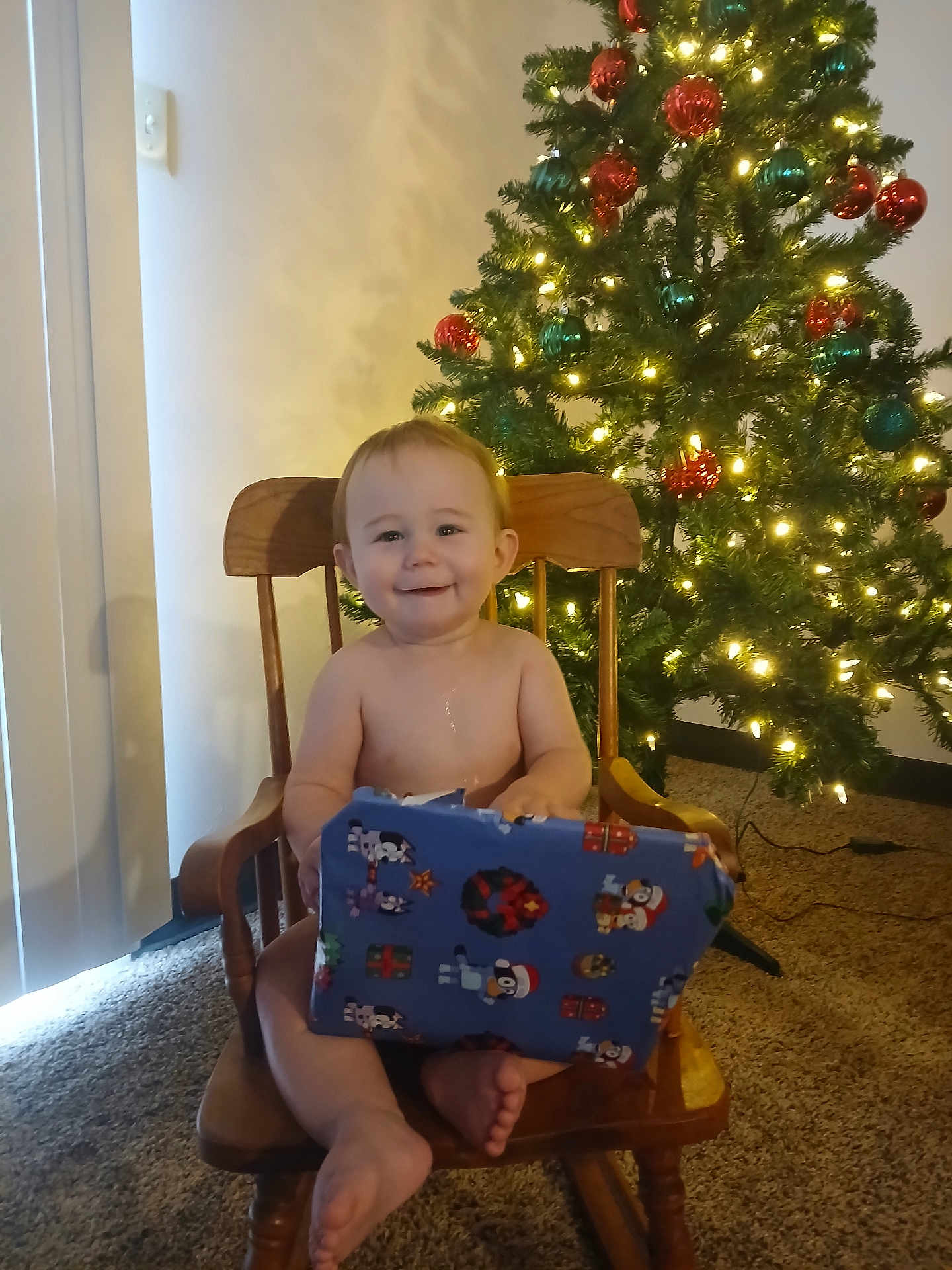 Tristan is registered to the contest to win money with this photo: toddler, child, smiling, rocking_chair, present, gift_wrap, christmas_tree, holiday_lights, carpet, indoor, festive, decorations, happy, home, celebration, seasonal, young_child, person, wooden_furniture, holiday