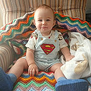 Tristan is registered to the contest to win money with this photo: baby, superman, blue_shoes, knitted_blanket, striped_chair, smile, cute, indoors, child, sitting, colorful, cozy, blanket, clothing, person, portrait, happy, infant, cute_baby, soft_light