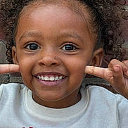 Kemani is registered to the contest to win money with this photo: child, smiling, face, curly_hair, hands, playful, close_up, brick_wall, white_shirt, red_text, joyful, person, portrait, happy, cute, young, skin, fingers, expression, outdoor