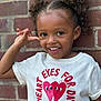 Kemani joined the competition — help win amazing prizes! child, smiling, curly_hair, tshirt, graphic_tee, brick_wall, playful, happy, portrait, casual_clothing, outdoor, young_child, cute, two_puffs, bright_eyes, joyful, person, fashion, expression, fun