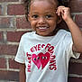 Kemani is registered to the contest to win money with this photo: child, smile, curly_hair, tshirt, heart_graphic, brick_wall, portrait, happy, outdoor, person, cute, young, fashion, casual, wall, standing, face, expression, fun, playful