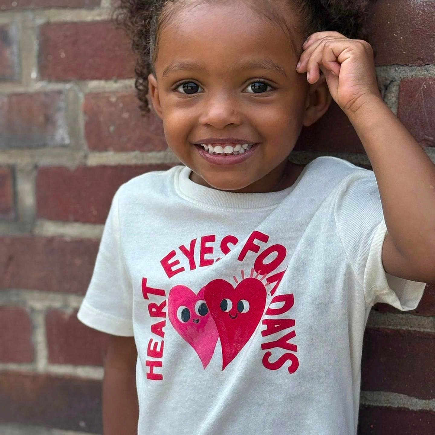Kemani is registered to the contest to win money with this photo: brick_wall, casual, child, curly_hair, cute, expression, face, fashion, fun, happy, heart_graphic, outdoor, person, playful, portrait, smile, standing, tshirt, wall, young