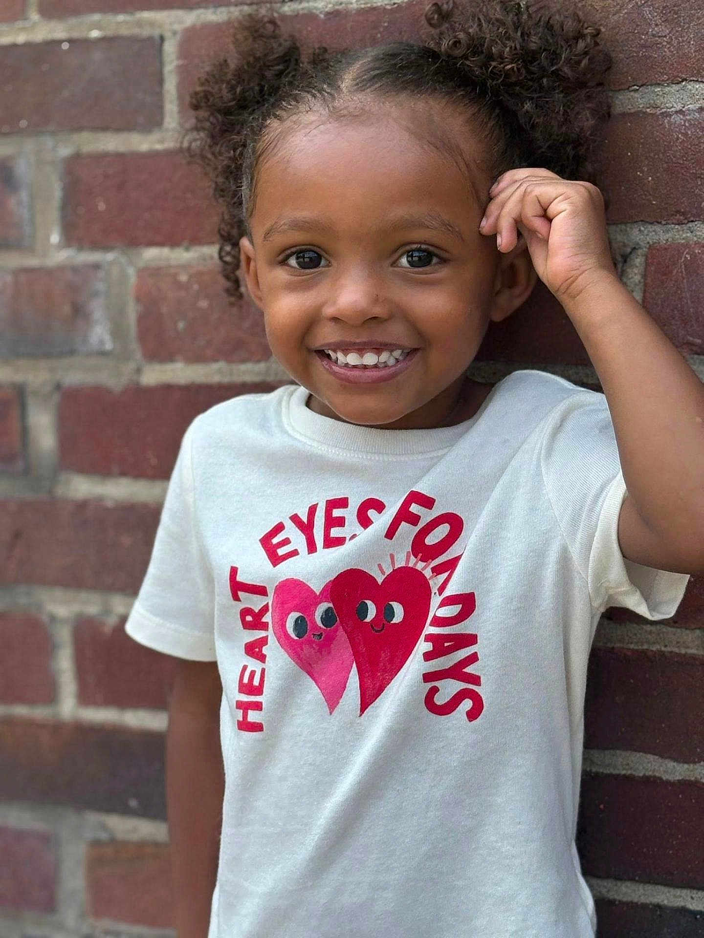 Kemani is registered to the contest to win money with this photo: child, smile, curly_hair, tshirt, heart_graphic, brick_wall, portrait, happy, outdoor, person, cute, young, fashion, casual, wall, standing, face, expression, fun, playful