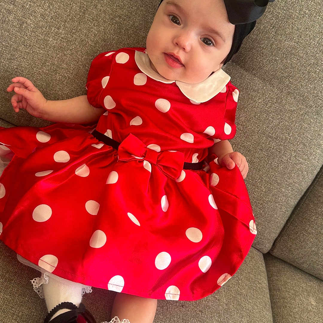Kaïly participe au concours pour gagner de l'argent avec cette photo : baby, child, red_dress, polka_dot, black_bow, headband, couch, sofa, smile, cute, infant, footwear, white_socks, portrait, indoors, relaxed, fashion, toddler, clothing, seat