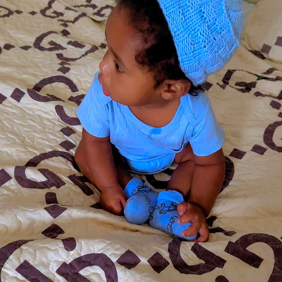 Jaden is registered to the contest to win money with this photo: baby, child, toddler, sitting, bed, bedroom, blue_outfit, knitted_hat, hat, portrait, profile, face, person, baby_socks, curly_hair, looking_sideways, indoor, bedding, patterned_blanket, photography