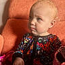 toddler, child, face, blonde_hair, colorful_clothing, robot_pattern, orange_chair, indoor, portrait, cute, expression, hand, holding, cup, blanket, cozy, seated, young_child, curious, soft_light