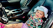 Addie is registered to the contest to win money with this photo: toddler, child, car_seat, leather_seat, hat, smiling, colorful_clothing, sandal, vehicle_interior, steering_wheel, window, person_outside, red_car, outdoor, daylight, happy, casual, seatbelt, portrait, relaxing