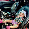 toddler, child, car_seat, leather_seat, hat, smiling, colorful_clothing, sandal, vehicle_interior, steering_wheel, window, person_outside, red_car, outdoor, daylight, happy, casual, seatbelt, portrait, relaxing