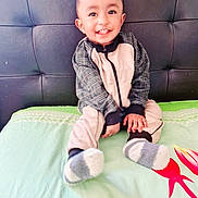 Ibrahim is registered to the contest to win money with this photo: child, toddler, smiling, socks, jacket, bed, pillow, headboard, happy, indoor, person, clothing, young_child, sitting, cozy, cute, portrait, home, comfortable, casual