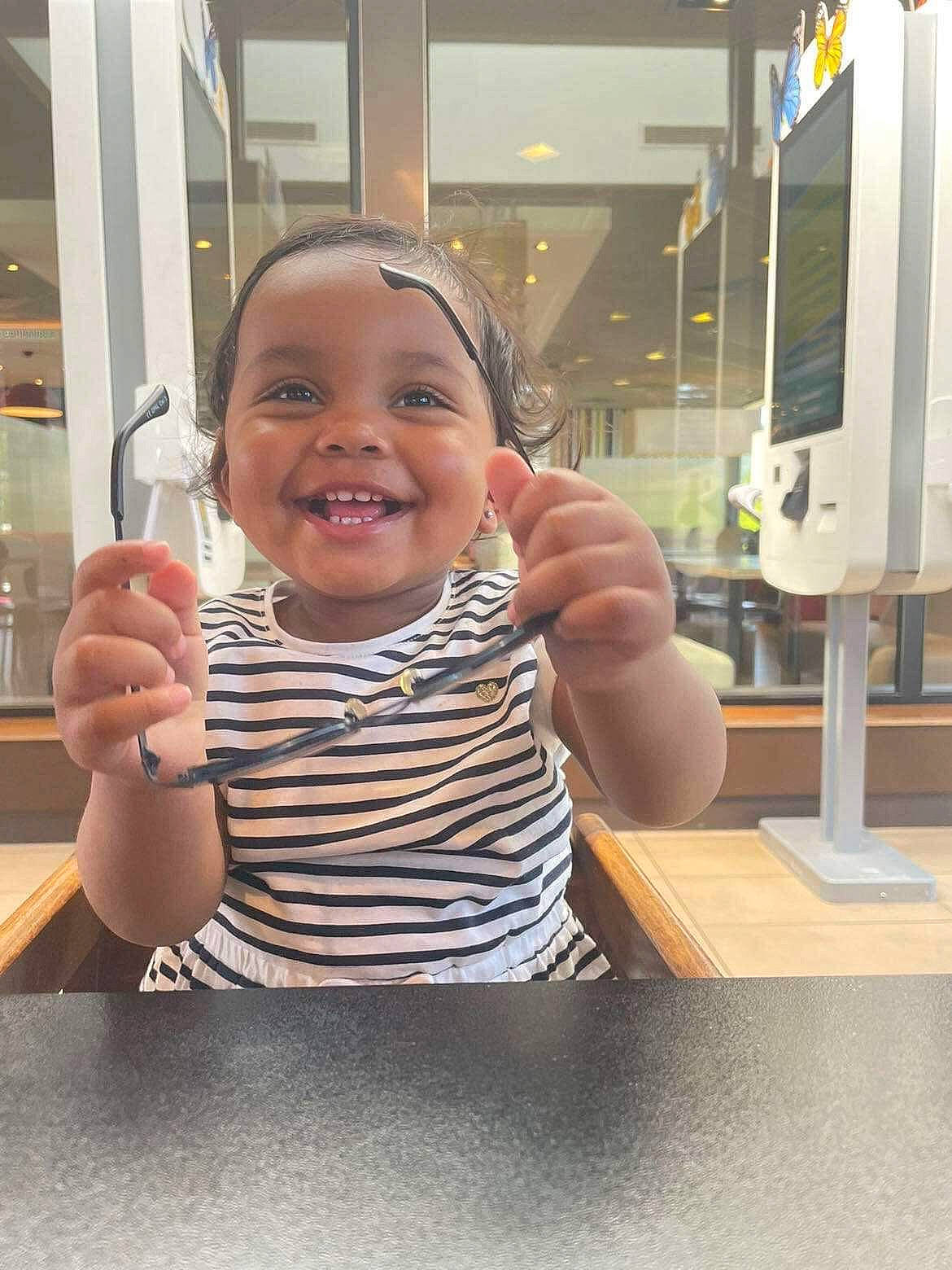 Maddie a rejoint le concours — aidez-le/la à gagner de superbes lots ! baby_toddler_clothing, child, finger, flooring, fun, gesture, hand, happy, joy, laugh, person, room, sitting, sleeve, smile, t_shirt, table, tableware, thumb, toddler