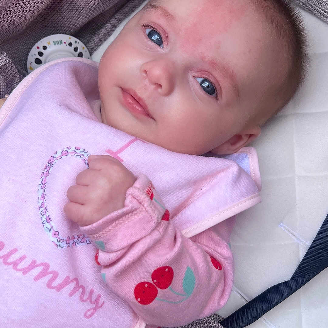 Mai joined the competition — help win amazing prizes! baby, bib, blanket, cherry_pattern, child, closeup, cushion, cute, expression, face, hand, indoor, infant, newborn, pacifier, person, pink_clothing, portrait, sleepwear, soft_light