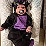 baby, child, costume, pacifier, purple, black, monster, cute, infant, lying_down, indoors, soft_lighting, cushion, playful, clothing, face, headgear, hand, footwear, person