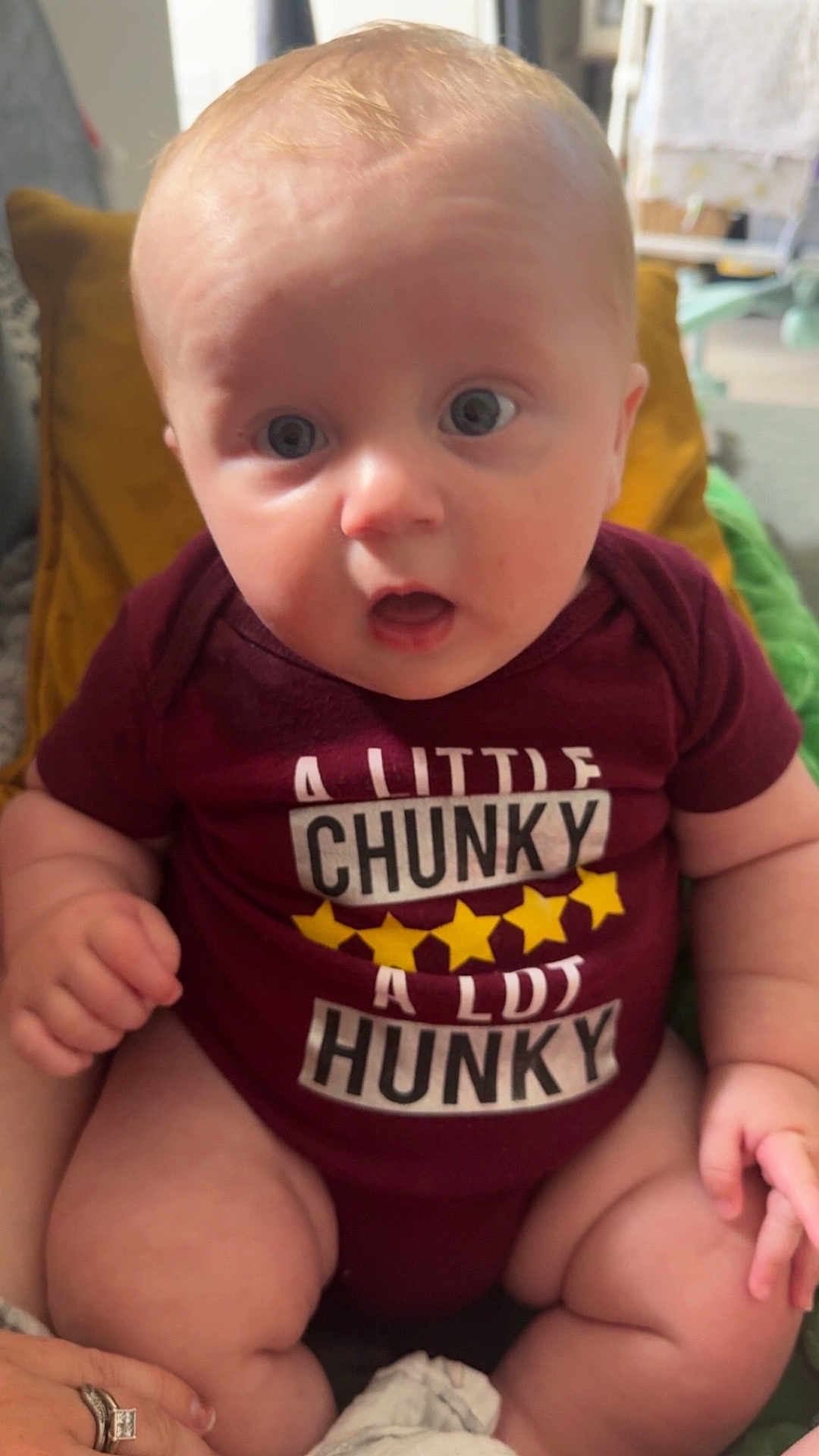 Dylan is registered to the contest to win money with this photo: baby, infant, child, onesie, maroon_clothing, chubby, sitting, indoor, face, wide_eyes, expression, hand, legs, pillow, blurred_background, home, cute, person, close_up, casual