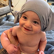 Staton is registered to the contest to win money with this photo: baby, child, infant, smiling, happy, portrait, gray_hat, diaper, indoor, home, blanket, adult_hand, tattoo, sitting, close_up, face, eyes, bare_skin, cozy, photography
