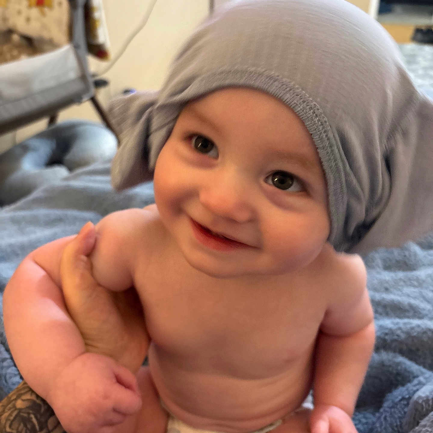 Staton is registered to the contest to win money with this photo: baby, child, infant, smiling, happy, portrait, gray_hat, diaper, indoor, home, blanket, adult_hand, tattoo, sitting, close_up, face, eyes, bare_skin, cozy, photography