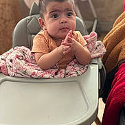 Alanï joined the competition — help win amazing prizes! baby, high_chair, blanket, infant, hands, clothing, face, person, indoor, curious_expression, pink, brown, wood_floor, foot, table, cozy, seated, young_child, cute, portrait
