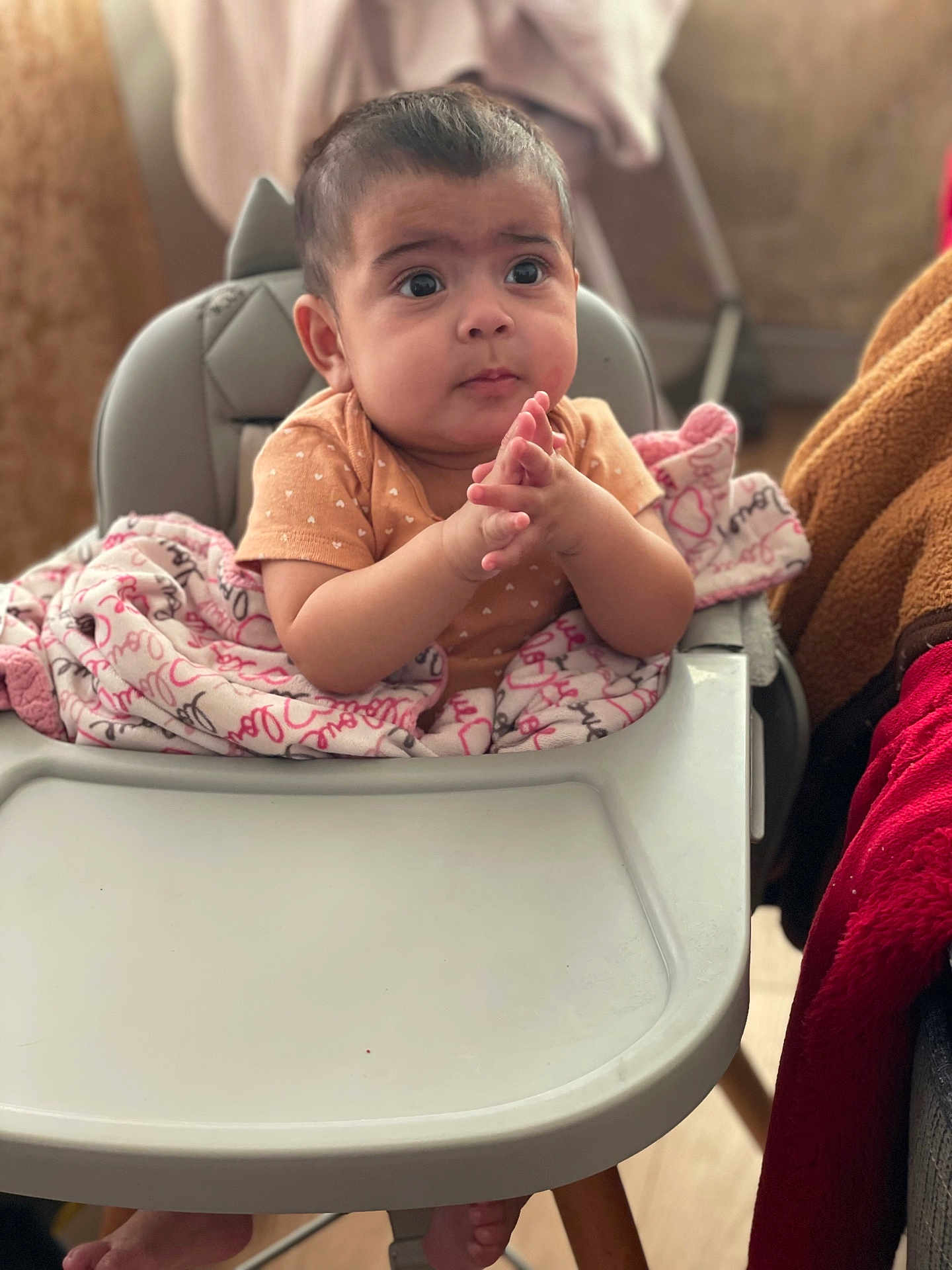 Alanï joined the competition — help win amazing prizes! baby, high_chair, blanket, infant, hands, clothing, face, person, indoor, curious_expression, pink, brown, wood_floor, foot, table, cozy, seated, young_child, cute, portrait