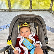 Rayan is registered to the contest to win money with this photo: baby, child, crown, star_wand, red_robe, blue_sash, stroller, smile, outdoor, deck, wooden_floor, fence, water, greenery, portrait, cute, happy, costume, person, infant