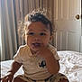 baby, child, curly_hair, smiling, sitting, bed, white_shirt, elephant_print, patterned_shorts, sock, indoor, curtains, door, natural_light, cute, playful, finger_in_mouth, portrait, person, cozy