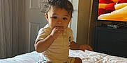 Zakar is registered to the contest to win money with this photo: toddler, child, bed, indoor, curly_hair, white_bedspread, sitting, hand_in_mouth, door, television, furniture, room, casual_clothing, shorts, socks, curious, young_child, portrait, home, cute
