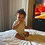 toddler, child, bed, indoor, curly_hair, white_bedspread, sitting, hand_in_mouth, door, television, furniture, room, casual_clothing, shorts, socks, curious, young_child, portrait, home, cute
