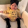 baby, infant, bow_headband, black_bow, onesie, text_on_clothing, smiling, cozy_chair, plush_chair, knee_high_socks, black_socks, yellow_outfit, knit_blanket, embroidered_text, portrait, indoor, chubby_cheeks, hands, legs, cute