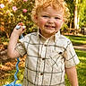 toddler, child, curly_hair, blue_eyes, smiling, plaid_shirt, outdoor, garden, flowers, greenery, sunlight, toy, knitted_toy, happy, portrait, grass, nature, cute, boy, casual_clothing