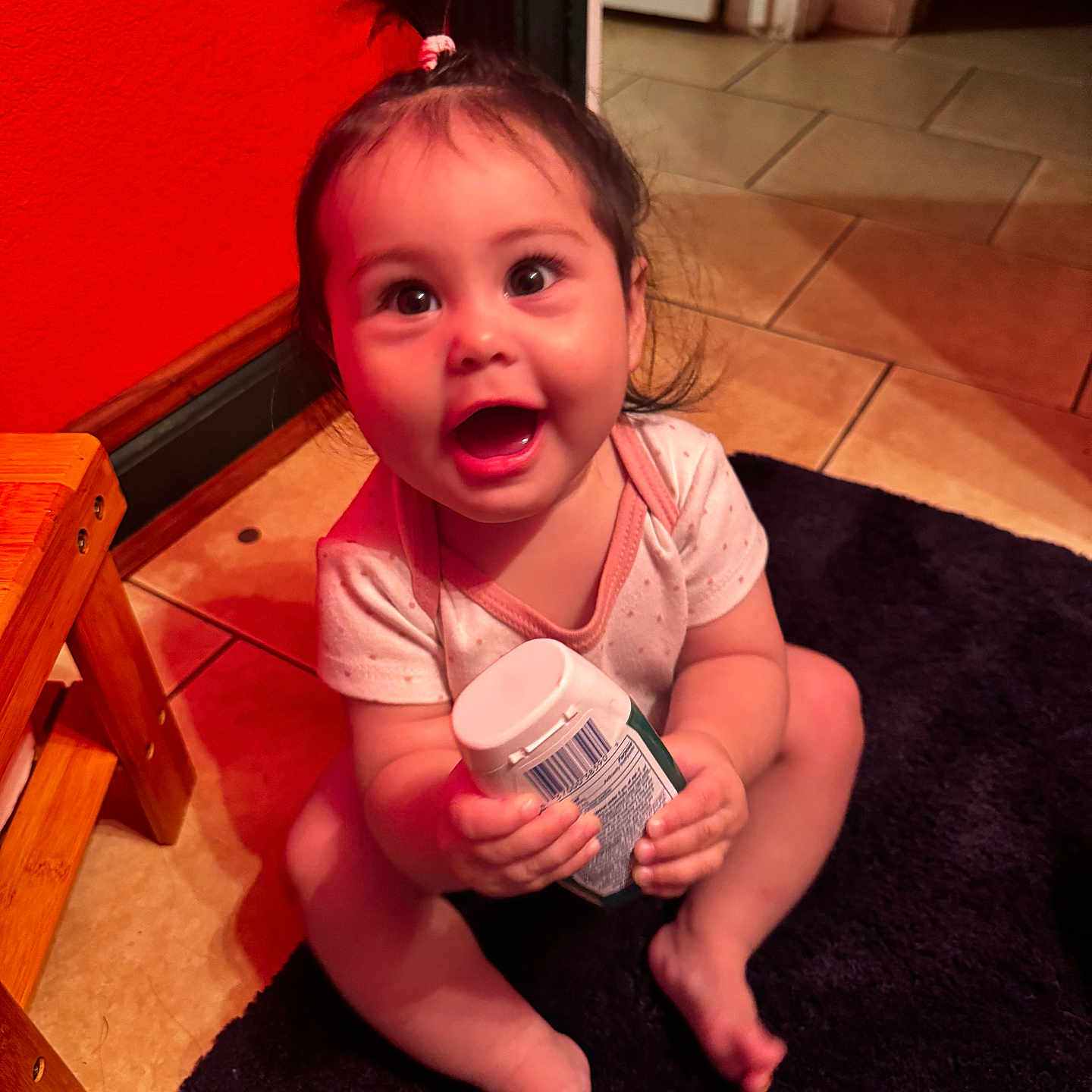 Alora is registered to the contest to win money with this photo: baby, bottle, bright, carpet, child, cute, expression, floor, foot, hair_tie, hand, happy, indoor, onesie, person, ponytail, sitting, smiling, tile, wooden_stool