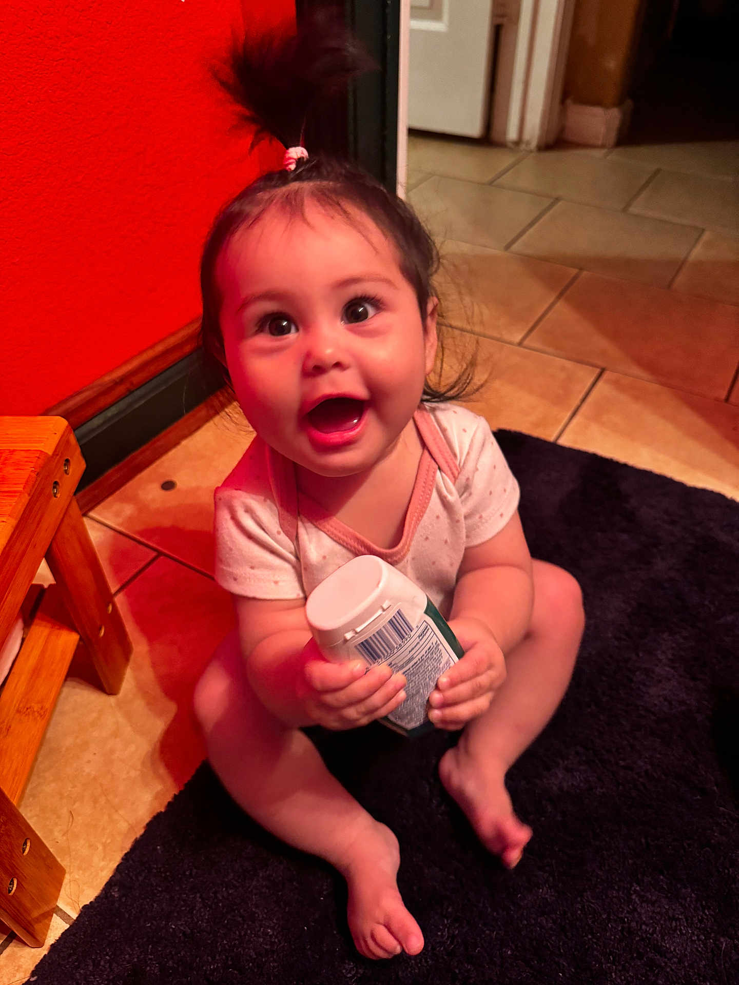 Alora is registered to the contest to win money with this photo: baby, child, indoor, smiling, happy, sitting, floor, carpet, tile, bottle, wooden_stool, hair_tie, ponytail, onesie, person, cute, bright, expression, hand, foot