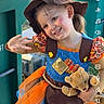 child, girl, smiling, costume, witch_hat, halloween, teddy_bear, peace_sign, outdoor, portrait, green_bins, waste_management, orange_dress, colorful_clothing, flower, face_paint, blonde_hair, happy, playful, photograph