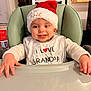 baby, child, high_chair, santa_hat, bib, food, messy, smile, indoor, holiday, festive, clothing, face, hands, table, chair, person, cute, expression, home