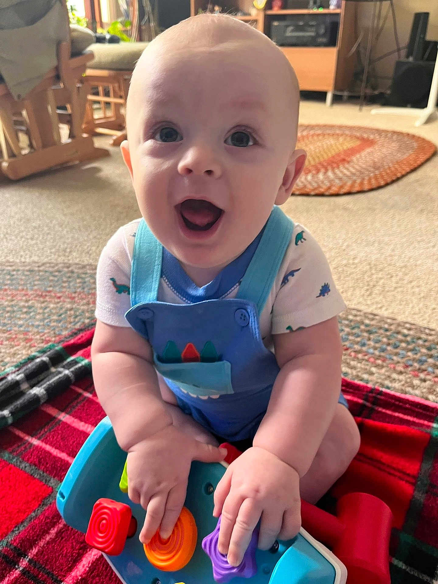 Tatum is registered to the contest to win money with this photo: baby, child, toy, blue_overalls, plaid_blanket, indoor, carpet, smiling, happy, infant, person, cute, play, hands, face, sitting, bright_eyes, home, colorful, clothing